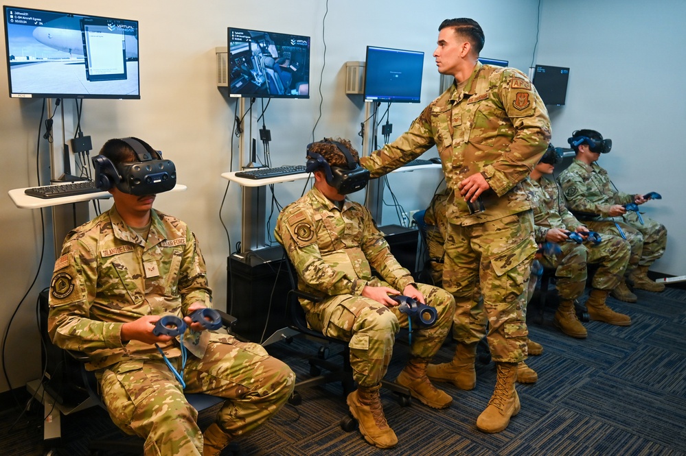 Military personnel in U.S. Air Force OCP (Operational Camouflage Pattern) uniforms are participating in a virtual reality training session. Several airmen are seated wearing VR headsets and holding motion controllers, while one standing individual appears to be an instructor assisting or supervising the trainees. Multiple monitors in the background display VR simulation feeds, including what appears to be aircraft-related training content.