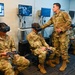 Military personnel in U.S. Air Force OCP (Operational Camouflage Pattern) uniforms are participating in a virtual reality training session. Several airmen are seated wearing VR headsets and holding motion controllers, while one standing individual appears to be an instructor assisting or supervising the trainees. Multiple monitors in the background display VR simulation feeds, including what appears to be aircraft-related training content.
