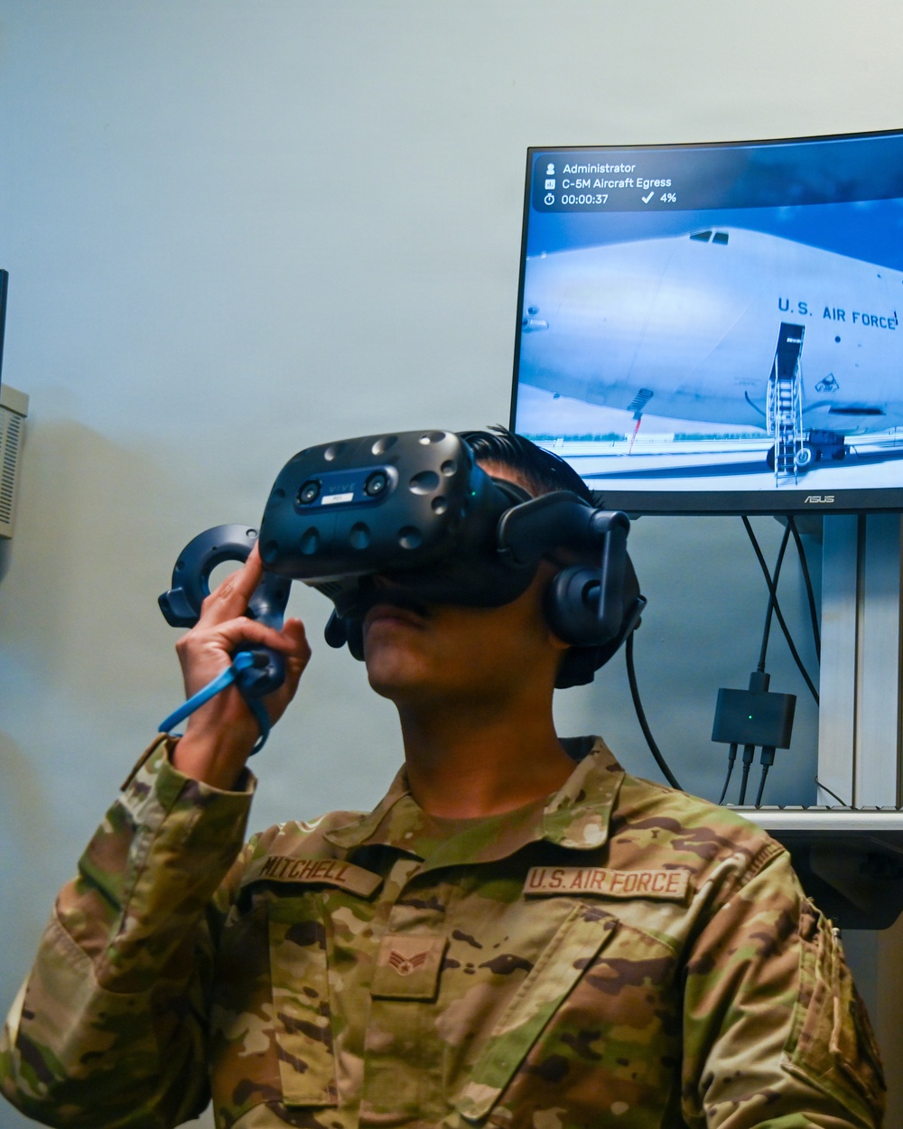A U.S. Air Force Airman named Mitchell is using a VR headset (HTC Vive) and hand controllers to participate in a virtual training simulation. The monitor behind them displays a C-5M Aircraft Egress training module, showing a U.S. Air Force aircraft exterior. The simulation is at 4% completion with a timer reading 00:00:37.
