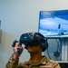 A U.S. Air Force Airman named Mitchell is using a VR headset (HTC Vive) and hand controllers to participate in a virtual training simulation. The monitor behind them displays a C-5M Aircraft Egress training module, showing a U.S. Air Force aircraft exterior. The simulation is at 4% completion with a timer reading 00:00:37.