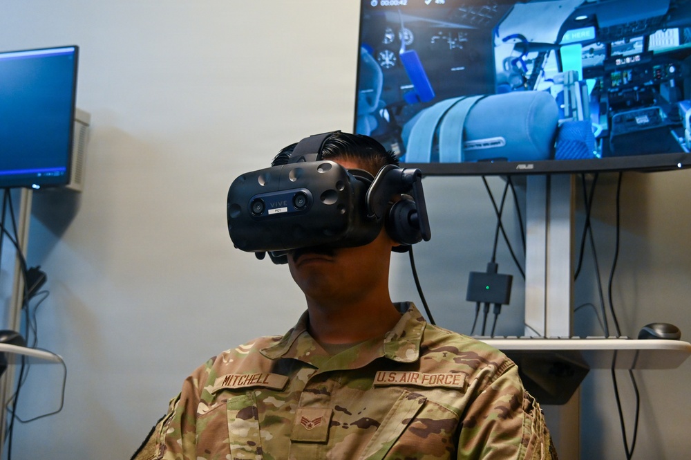 A U.S. Air Force service member named Mitchell is wearing an HTC Vive Pro VR headset, appearing to engage in a virtual reality training simulation. Behind them, an ASUS monitor displays what appears to be a cockpit or aircraft interior simulation environment. The setting appears to be a military training facility equipped with VR technology.