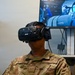 A U.S. Air Force service member named Mitchell is wearing an HTC Vive Pro VR headset, appearing to engage in a virtual reality training simulation. Behind them, an ASUS monitor displays what appears to be a cockpit or aircraft interior simulation environment. The setting appears to be a military training facility equipped with VR technology.