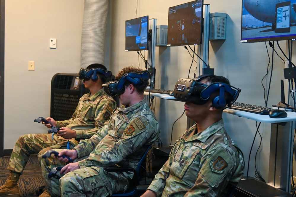 Three military personnel in OCP (Operational Camouflage Pattern) uniforms are seated and using virtual reality headsets in what appears to be a VR training facility. They are holding VR controllers and wearing HTC Vive-style headsets while multiple monitors on the wall display their virtual environments. This appears to be a military simulation or training exercise using immersive VR technology.