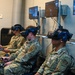 Three military personnel in OCP (Operational Camouflage Pattern) uniforms are seated and using virtual reality headsets in what appears to be a VR training facility. They are holding VR controllers and wearing HTC Vive-style headsets while multiple monitors on the wall display their virtual environments. This appears to be a military simulation or training exercise using immersive VR technology.