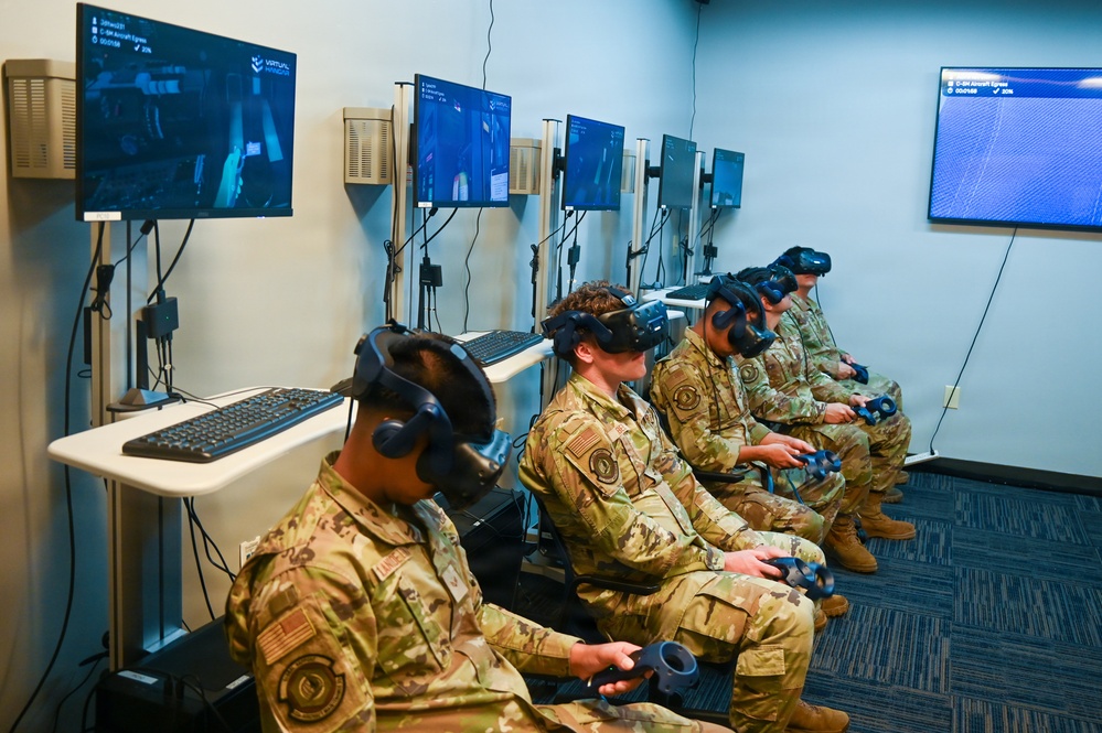 Four military personnel in camouflage uniforms are seated in a row using VR headsets and handheld controllers in what appears to be a dedicated virtual reality training facility. Multiple monitors mounted on the wall display the VR simulation content being experienced by each trainee. The screen text visible on monitors references 'C-5M Aircraft Egress,' indicating this is likely an aircraft emergency egress training simulation.