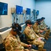 Four military personnel in camouflage uniforms are seated in a row using VR headsets and handheld controllers in what appears to be a dedicated virtual reality training facility. Multiple monitors mounted on the wall display the VR simulation content being experienced by each trainee. The screen text visible on monitors references 'C-5M Aircraft Egress,' indicating this is likely an aircraft emergency egress training simulation.