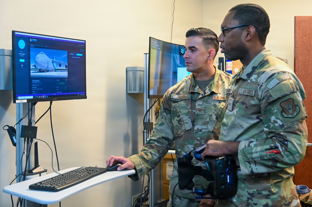 Two U.S. Air Force personnel in OCP (Operational Camouflage Pattern) uniforms are examining a computer workstation displaying what appears to be a large military transport aircraft (likely a C-17 or C-5) on screen. One airman is operating a keyboard and mouse while the other holds what appears to be a VR headset or controller, suggesting they are engaged in a virtual reality or simulation training environment. The setting appears to be an indoor training or technology demonstration room.