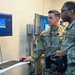 Two U.S. Air Force personnel in OCP (Operational Camouflage Pattern) uniforms are examining a computer workstation displaying what appears to be a large military transport aircraft (likely a C-17 or C-5) on screen. One airman is operating a keyboard and mouse while the other holds what appears to be a VR headset or controller, suggesting they are engaged in a virtual reality or simulation training environment. The setting appears to be an indoor training or technology demonstration room.