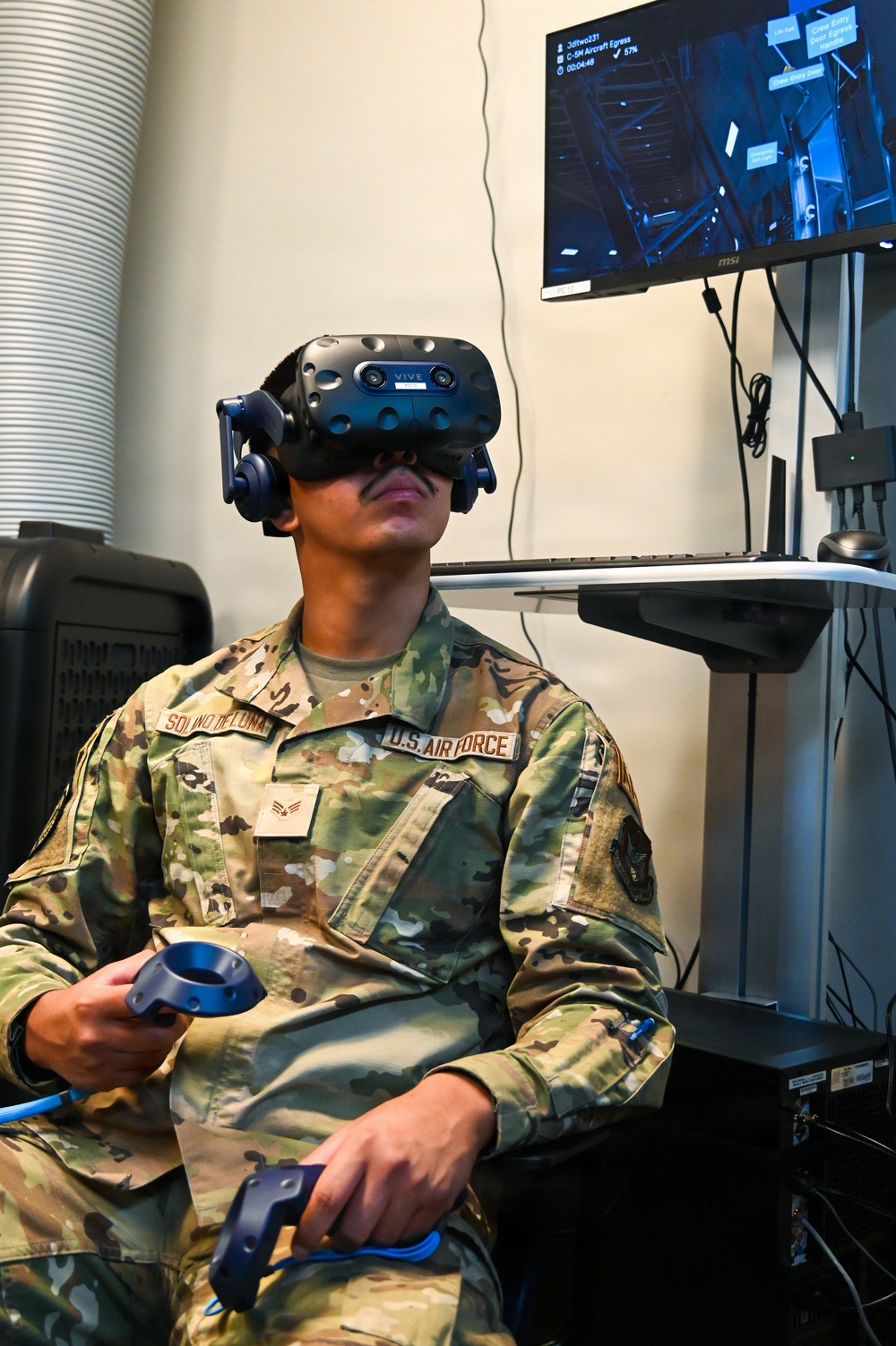 A U.S. Air Force airman wearing an HTC Vive VR headset is seated and engaged in a virtual reality training simulation. The airman, identified by name tape as 'Solano DeLuna' with a Senior Airman rank insignia, holds two VR controllers while a monitor behind displays the simulation screen labeled 'C-5M Aircraft Egress.' This appears to be a military training exercise using immersive VR technology.