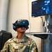 A U.S. Air Force airman wearing an HTC Vive VR headset is seated and engaged in a virtual reality training simulation. The airman, identified by name tape as 'Solano DeLuna' with a Senior Airman rank insignia, holds two VR controllers while a monitor behind displays the simulation screen labeled 'C-5M Aircraft Egress.' This appears to be a military training exercise using immersive VR technology.