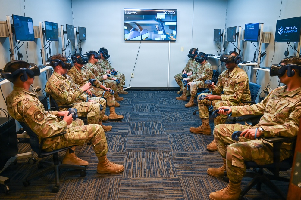 A group of military personnel in camouflage uniforms are seated in a semicircular arrangement in a dedicated VR training room, all wearing virtual reality headsets and holding VR controllers. The room is equipped with multiple monitor stations along the walls and a central display screen showing 'Virtual Hangar' software, suggesting aircraft maintenance or aviation training. This appears to be a formal military VR simulation training session.