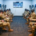 A group of military personnel in camouflage uniforms are seated in a semicircular arrangement in a dedicated VR training room, all wearing virtual reality headsets and holding VR controllers. The room is equipped with multiple monitor stations along the walls and a central display screen showing 'Virtual Hangar' software, suggesting aircraft maintenance or aviation training. This appears to be a formal military VR simulation training session.
