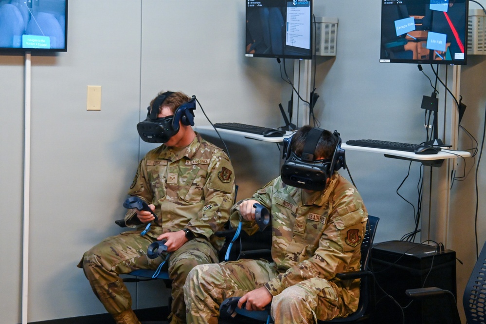 Two U.S. Air Force personnel in OCP (Operational Camouflage Pattern) uniforms are seated and using VR (Virtual Reality) headsets with hand controllers in what appears to be a training facility. Behind them, multiple monitors mounted on the wall display what appears to be the VR simulation content they are experiencing. This appears to be a military VR training session.