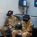Two U.S. Air Force personnel in OCP (Operational Camouflage Pattern) uniforms are seated and using VR (Virtual Reality) headsets with hand controllers in what appears to be a training facility. Behind them, multiple monitors mounted on the wall display what appears to be the VR simulation content they are experiencing. This appears to be a military VR training session.