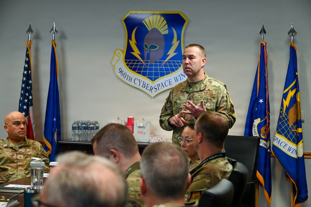 A U.S. Air Force officer is addressing a group of military personnel in a conference room setting. The meeting is taking place at the 960th Cyberspace Wing, as indicated by the unit emblem prominently displayed on the wall. The speaker is standing and gesturing while uniformed personnel are seated around a conference table.