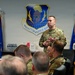 A U.S. Air Force officer is addressing a group of military personnel in a conference room setting. The meeting is taking place at the 960th Cyberspace Wing, as indicated by the unit emblem prominently displayed on the wall. The speaker is standing and gesturing while uniformed personnel are seated around a conference table.