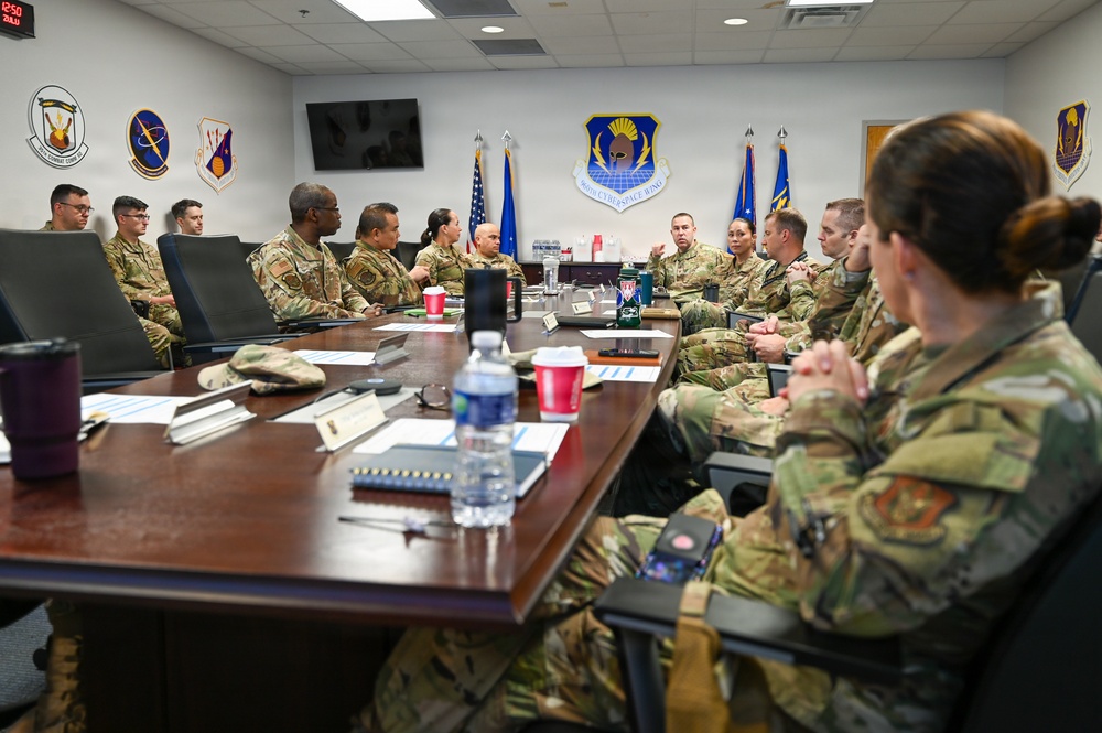 A group of U.S. Air Force personnel in OCP (Operational Camouflage Pattern) uniforms are seated around a large conference table in what appears to be an official briefing or meeting room. The room displays multiple unit emblems on the walls, including a central shield identified as the 960th Cyberspace Wing, along with U.S. and Air Force flags. The meeting appears to involve active discussion among the participants.