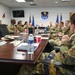 A group of U.S. Air Force personnel in OCP (Operational Camouflage Pattern) uniforms are seated around a large conference table in what appears to be an official briefing or meeting room. The room displays multiple unit emblems on the walls, including a central shield identified as the 960th Cyberspace Wing, along with U.S. and Air Force flags. The meeting appears to involve active discussion among the participants.