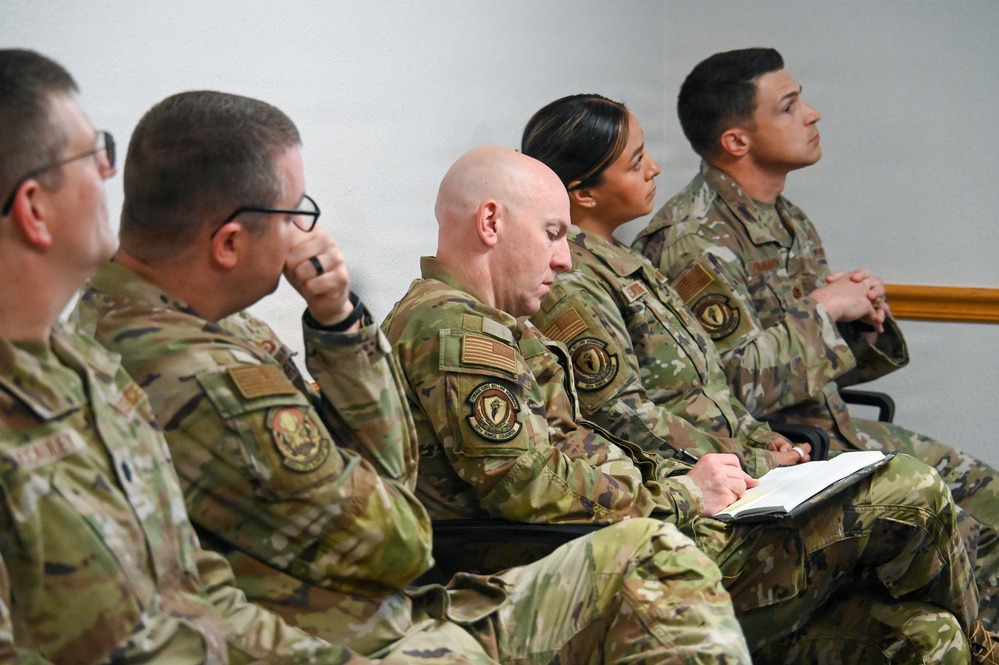 A group of five military personnel in OCP (Operational Camouflage Pattern) uniforms are seated in a row, appearing to attend a briefing or training session. They display varying levels of engagement, with one individual taking notes in a notebook while others listen attentively. All personnel wear U.S. flag patches and unit patches on their sleeves, consistent with U.S. Air Force or Space Force uniforms.