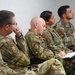 A group of five military personnel in OCP (Operational Camouflage Pattern) uniforms are seated in a row, appearing to attend a briefing or training session. They display varying levels of engagement, with one individual taking notes in a notebook while others listen attentively. All personnel wear U.S. flag patches and unit patches on their sleeves, consistent with U.S. Air Force or Space Force uniforms.