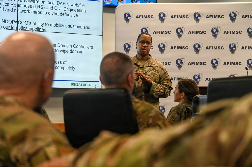 A military briefing is taking place at an AFIMSC (Air Force Installation and Mission Support Center) facility. A uniformed service member is presenting to an audience of other military personnel, with a presentation slide visible on a screen marked 'UNCLASSIFIED'. The backdrop features repeated AFIMSC branding with a globe logo.