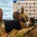 A military briefing is taking place at an AFIMSC (Air Force Installation and Mission Support Center) facility. A uniformed service member is presenting to an audience of other military personnel, with a presentation slide visible on a screen marked 'UNCLASSIFIED'. The backdrop features repeated AFIMSC branding with a globe logo.