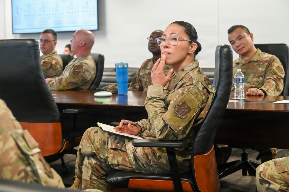 A group of military personnel in OCP (Operational Camouflage Pattern) uniforms are seated around a conference table during what appears to be a briefing or meeting. A female service member in the foreground is holding a notebook and pen while attentively listening, with her hand raised to her chin in a contemplative pose. A presentation screen is visible in the background.