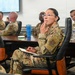 A group of military personnel in OCP (Operational Camouflage Pattern) uniforms are seated around a conference table during what appears to be a briefing or meeting. A female service member in the foreground is holding a notebook and pen while attentively listening, with her hand raised to her chin in a contemplative pose. A presentation screen is visible in the background.