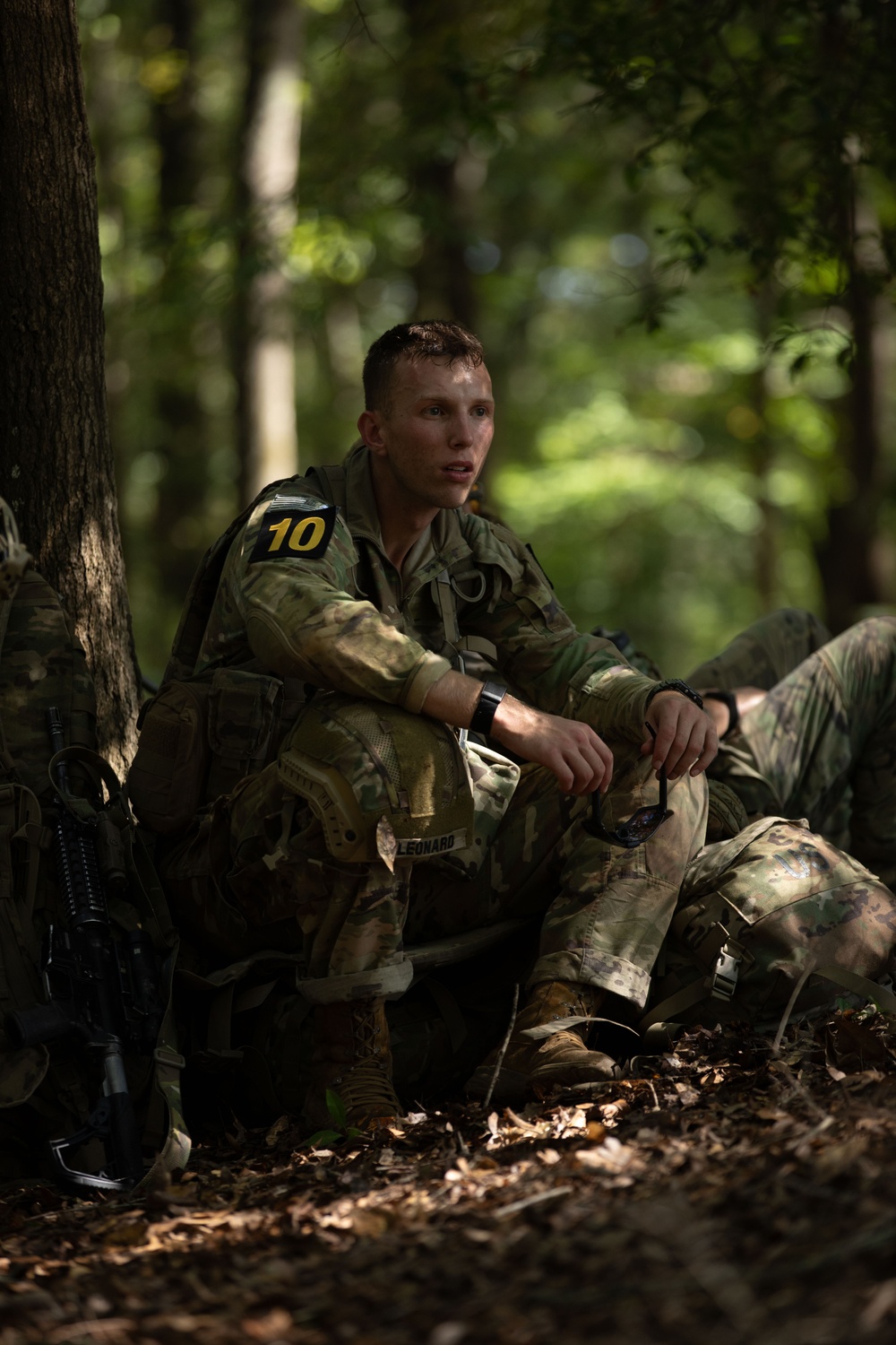 A young soldier in camouflage uniform is resting against a tree in a wooded area, appearing fatigued with sweat visible on his face. He wears a number '10' patch on his shoulder and a name tape reading 'LEONARD' on his chest. The setting suggests a military training exercise or field operation.