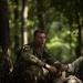 A young soldier in camouflage uniform is resting against a tree in a wooded area, appearing fatigued with sweat visible on his face. He wears a number '10' patch on his shoulder and a name tape reading 'LEONARD' on his chest. The setting suggests a military training exercise or field operation.