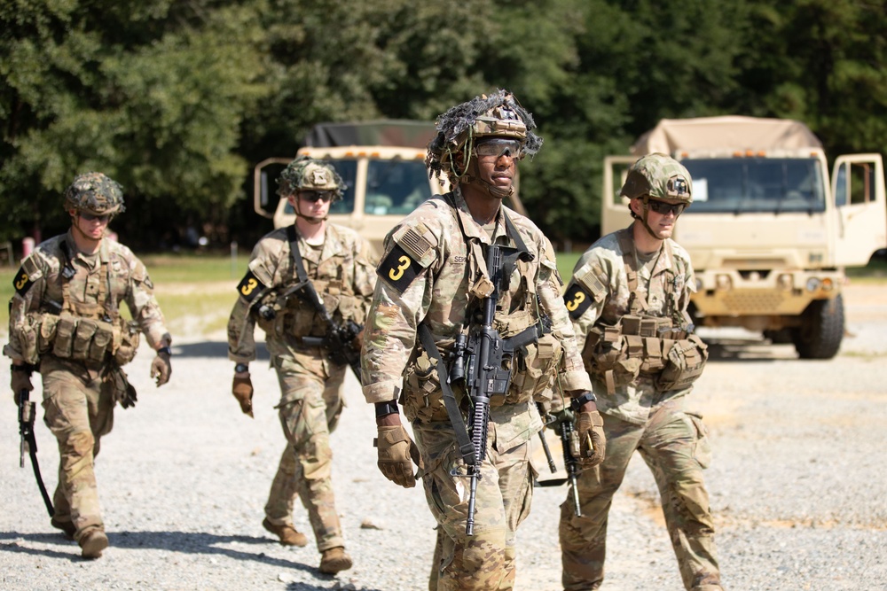Four soldiers in U.S. Army OCP (Operational Camouflage Pattern) uniforms are walking along a gravel path during what appears to be a training exercise. All personnel are wearing tactical gear, helmets, and carrying M4-style rifles, with black armbands marked with the number '3' visible on each soldier. Military vehicles, consistent with LMTV or FMTV trucks, are visible in the background.