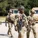 Four soldiers in U.S. Army OCP (Operational Camouflage Pattern) uniforms are walking along a gravel path during what appears to be a training exercise. All personnel are wearing tactical gear, helmets, and carrying M4-style rifles, with black armbands marked with the number '3' visible on each soldier. Military vehicles, consistent with LMTV or FMTV trucks, are visible in the background.
