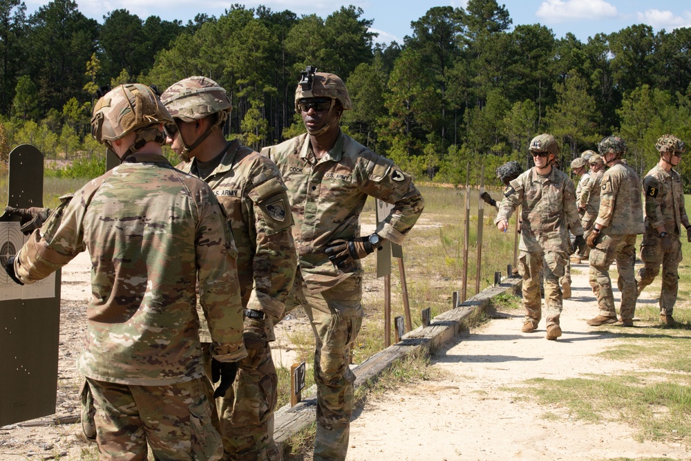 A group of U.S. Army soldiers are gathered at an outdoor firing range, appearing to conduct marksmanship training or qualification. A soldier identified by name tape as 'COOK' is centrally positioned and appears to be instructing or debriefing other soldiers near a shooting target. Additional soldiers are visible in the background along the range lane pathway.