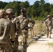 A group of U.S. Army soldiers are gathered at an outdoor firing range, appearing to conduct marksmanship training or qualification. A soldier identified by name tape as 'COOK' is centrally positioned and appears to be instructing or debriefing other soldiers near a shooting target. Additional soldiers are visible in the background along the range lane pathway.
