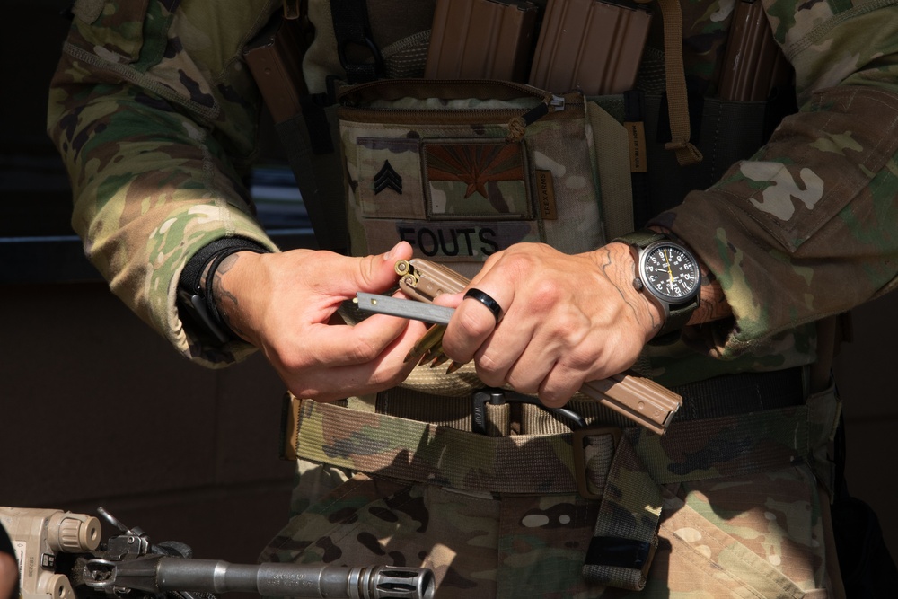 A soldier wearing OCP (Operational Camouflage Pattern) uniform and plate carrier is handling what appears to be a stripper clip with ammunition. The name tape on the plate carrier reads 'FOUTS' and a rank patch (Sergeant chevrons) is visible along with a state flag patch. The soldier is wearing a black analog watch and a ring on their right hand.