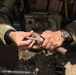 A soldier wearing OCP (Operational Camouflage Pattern) uniform and plate carrier is handling what appears to be a stripper clip with ammunition. The name tape on the plate carrier reads 'FOUTS' and a rank patch (Sergeant chevrons) is visible along with a state flag patch. The soldier is wearing a black analog watch and a ring on their right hand.
