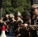 A group of five military personnel in MultiCam/OCP camouflage uniforms and tactical helmets are gathered around a table, appearing to review documents or conduct a briefing. The soldiers are equipped with tactical gear, body armor, and communications equipment. The setting appears to be an outdoor covered area near a building, likely during a training exercise or operational planning session.