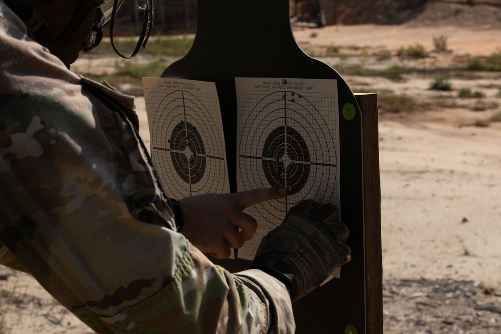 A military service member in camouflage uniform and tactical gloves is examining two paper shooting targets mounted on a dark silhouette target stand at an outdoor firing range. The person appears to be pointing at bullet holes on the right target to assess shot placement and accuracy. The setting is a dry, sandy outdoor range environment.