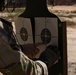 A military service member in camouflage uniform and tactical gloves is examining two paper shooting targets mounted on a dark silhouette target stand at an outdoor firing range. The person appears to be pointing at bullet holes on the right target to assess shot placement and accuracy. The setting is a dry, sandy outdoor range environment.