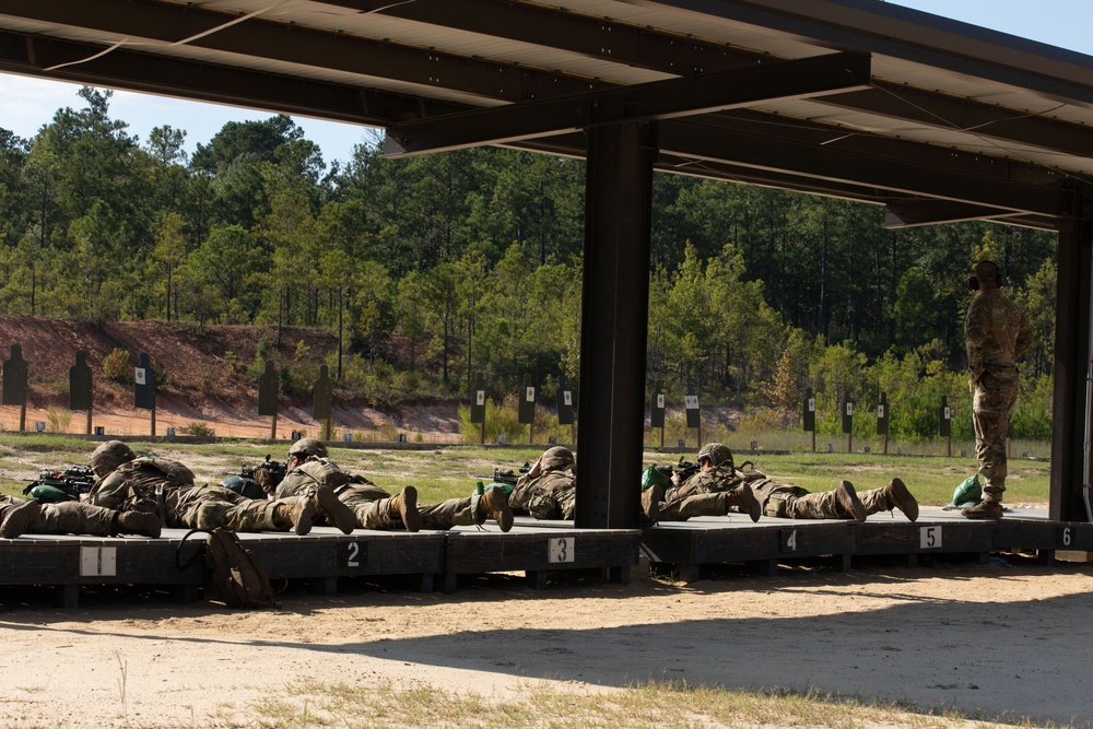 Military personnel are conducting a rifle qualification or marksmanship training exercise at an outdoor shooting range. Multiple soldiers are in the prone position on numbered firing platforms (lanes 1-6) aiming downrange at target silhouettes. A standing individual, likely a range safety officer or instructor, observes from the right side of the range.