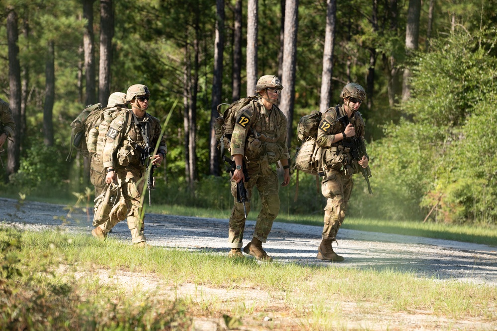 Three military personnel in camouflage uniforms are walking along a gravel road through a forested training area. All individuals are wearing full combat gear including helmets, body armor, rucksacks, and carrying rifles. Each soldier displays a yellow number '12' patch, suggesting a unit identifier or training exercise designation.