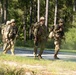 Three military personnel in camouflage uniforms are walking along a gravel road through a forested training area. All individuals are wearing full combat gear including helmets, body armor, rucksacks, and carrying rifles. Each soldier displays a yellow number '12' patch, suggesting a unit identifier or training exercise designation.