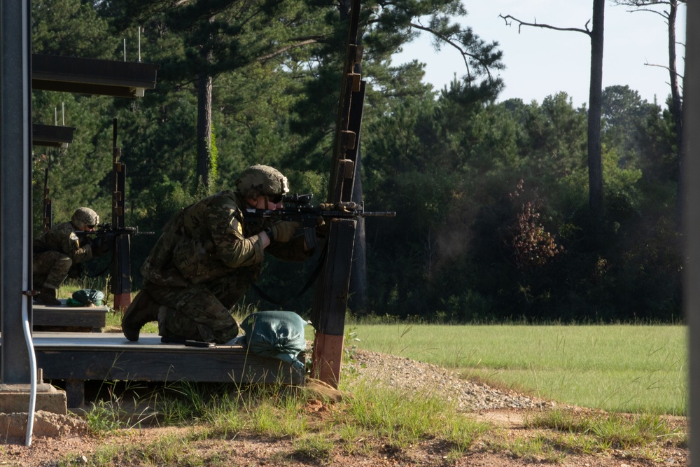 Two military personnel in camouflage uniforms and helmets are conducting a live-fire shooting exercise at what appears to be a military training range. The foreground soldier is in a kneeling position, aiming a rifle around a steel post barrier, with visible smoke/gas near the muzzle indicating recent discharge. The setting is an outdoor range with a grassy field downrange and pine trees in the background.