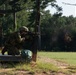 Two military personnel in camouflage uniforms and helmets are conducting a live-fire shooting exercise at what appears to be a military training range. The foreground soldier is in a kneeling position, aiming a rifle around a steel post barrier, with visible smoke/gas near the muzzle indicating recent discharge. The setting is an outdoor range with a grassy field downrange and pine trees in the background.