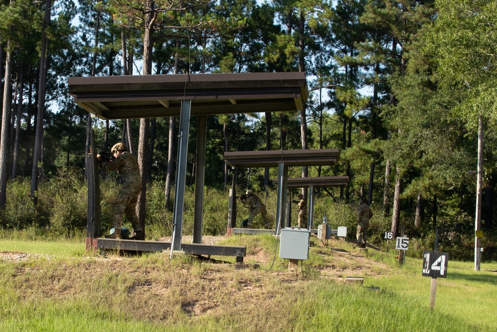 The image shows military personnel conducting firearms training at an outdoor shooting range with numbered firing positions. Multiple soldiers in camouflage uniforms are positioned under covered firing bays, appearing to engage targets downrange. The range is set in a wooded area with pine trees and features numbered lane markers visible in the foreground.
