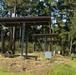 The image shows military personnel conducting firearms training at an outdoor shooting range with numbered firing positions. Multiple soldiers in camouflage uniforms are positioned under covered firing bays, appearing to engage targets downrange. The range is set in a wooded area with pine trees and features numbered lane markers visible in the foreground.