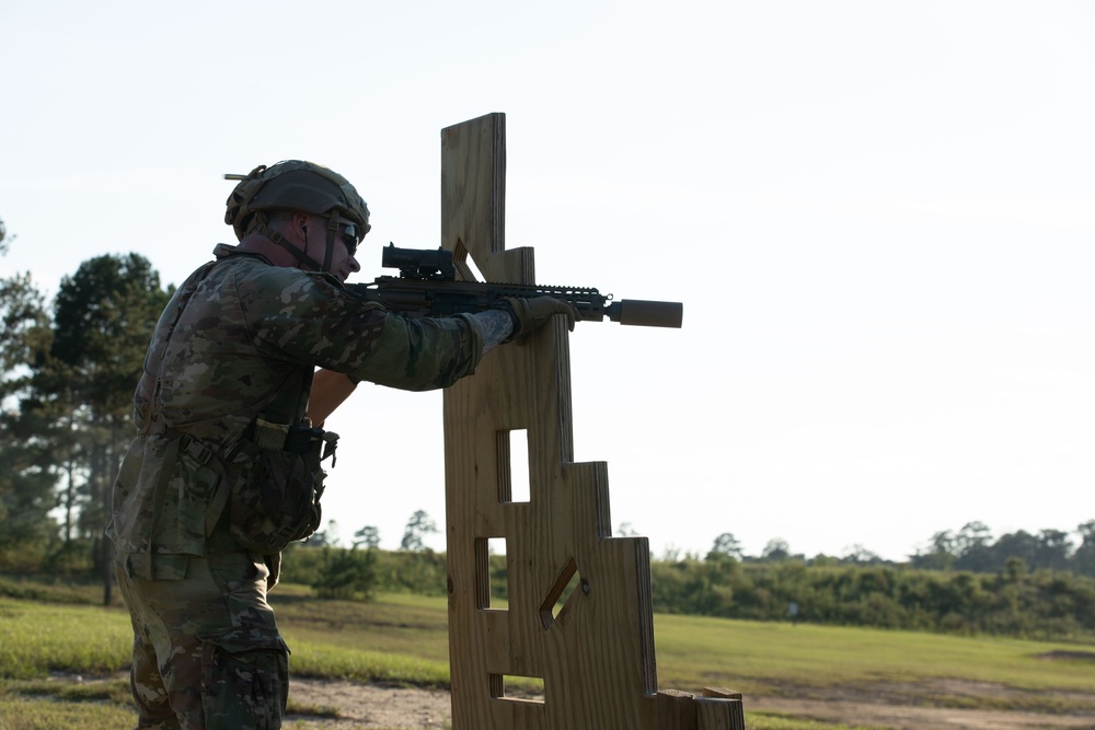 A soldier in full combat gear is conducting a shooting exercise at what appears to be a military training range. The individual is using a wooden barricade structure as cover while aiming a rifle equipped with an optic and suppressor. The setting is an outdoor range with open grassy terrain and tree line in the background.