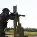 A soldier in full combat gear is conducting a shooting exercise at what appears to be a military training range. The individual is using a wooden barricade structure as cover while aiming a rifle equipped with an optic and suppressor. The setting is an outdoor range with open grassy terrain and tree line in the background.