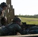 Two U.S. Army soldiers are at a firing range, with one soldier in the prone position aiming a rifle downrange while a second soldier stands behind observing and supervising. The standing soldier wears hearing protection and sunglasses, and displays a 10th Mountain Division patch on his left shoulder. The setting appears to be an outdoor military training range with sandbags, a wooden barricade structure, and an open grassy field in the background.