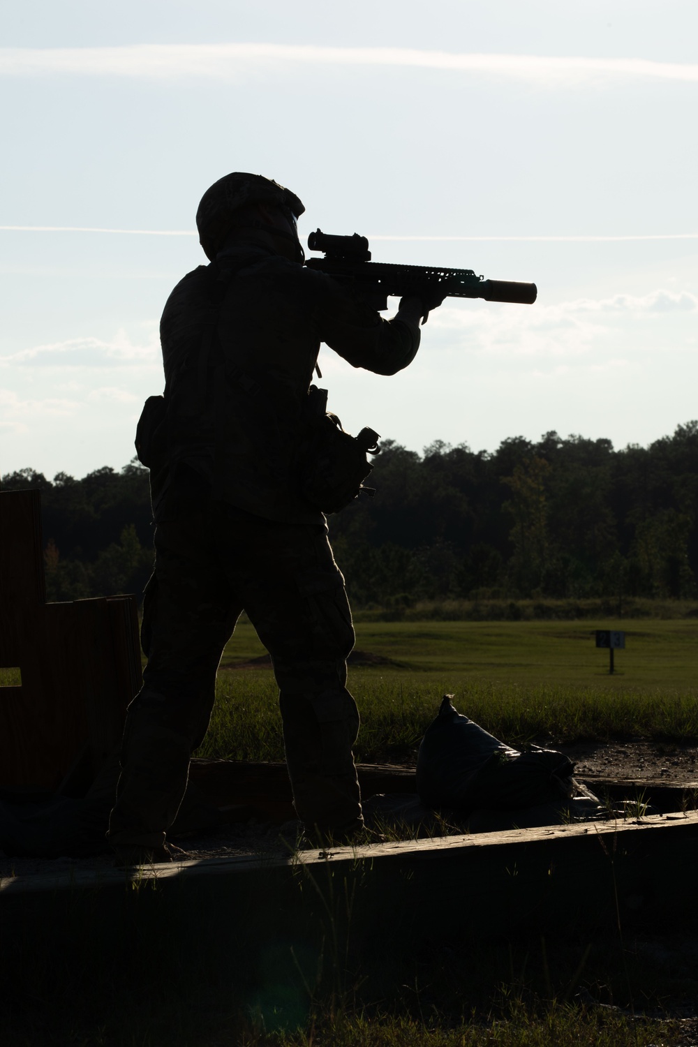 A silhouetted figure in full tactical gear is aiming a rifle equipped with a suppressor and optic sight at an outdoor shooting range. The backlit composition renders the subject entirely in shadow against a bright sky and open grassy field. The setting appears to be a military or law enforcement training range based on the infrastructure and equipment visible.